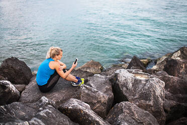 Young sporty woman runner with earphones and smartphone sitting on rocks by sea, listening to music. - HPIF29911