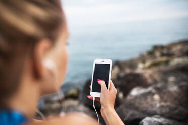 Unerkennbare junge sportliche Läuferin mit Kopfhörern und Smartphone auf einem Felsen am Meer sitzend, Musik hörend, Nahaufnahme. - HPIF29912