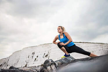 Young sporty woman runner with earphones standing outside on the rocks by beach in nature, listening to music and stretching. Copy space. - HPIF29919