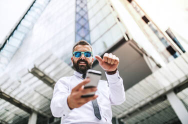 A low-angle view of hipster businessman with sunglasses and smartphone standing on the street in London, taking selfie. Copy space. - HPIF29946