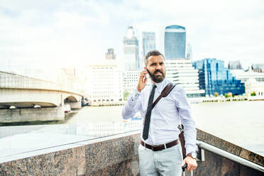 Hipster-Geschäftsmann mit Smartphone und Koffer, der an der Themse in London steht und telefoniert. Kopierraum. - HPIF29954