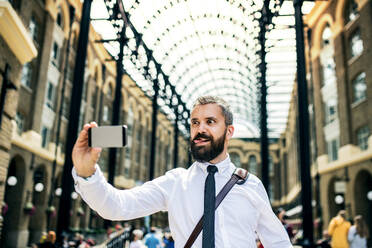 Hipster businessman with smartphone standing on the trian station in London, grimacing and taking selfie. - HPIF29957