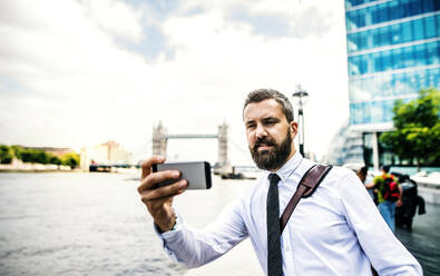 Hipster businessman with smartphone standing by the river Thames in London, taking selfie. Copy space. - HPIF29962