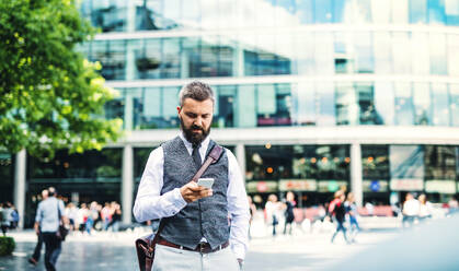 Hipster businessman with smartphone standing on the street in city, text messaging. - HPIF29964