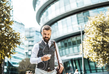 Hipster-Geschäftsmann mit Smartphone und Kopfhörern, der auf der Straße in London spazieren geht und Musik hört. - HPIF29972