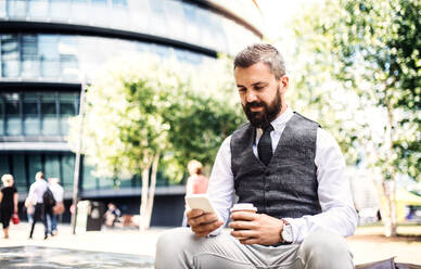 Seriöser Hipster-Geschäftsmann mit Smartphone und Kaffeetasse im Freien sitzend in der Stadt, Textnachrichten schreibend. - HPIF29975