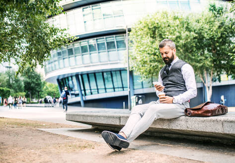 Seriöser Hipster-Geschäftsmann mit Smartphone, Laptoptasche und Kaffeetasse im Freien sitzend in der Stadt, Textnachrichten schreibend. - HPIF29976