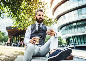 Hipster-Geschäftsmann mit Kaffee und Smartphone sitzt auf einer Bank auf der Straße in London und schreibt SMS. - HPIF29977