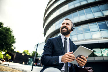 Hipster-Geschäftsmann mit Kopfhörern und Tablet auf einer Bank auf der Straße in London sitzend. Raum kopieren. - HPIF29979