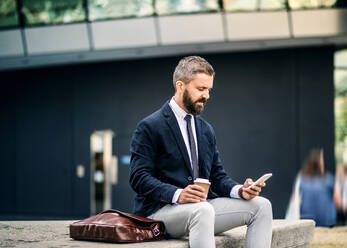 Seriöser Hipster-Geschäftsmann mit Smartphone, Laptoptasche und Kaffeetasse im Freien sitzend in der Stadt, Textnachrichten schreibend. - HPIF29981