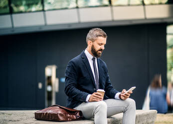 Serious hipster businessman with smartphone, laptop bag and coffee cup sitting outdoors in the city, text messaging. - HPIF29981