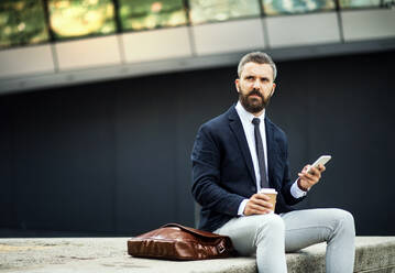 Serious hipster businessman with smartphone, laptop bag and coffee cup sitting outdoors in the city, text messaging. - HPIF29982