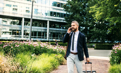 Hipster-Geschäftsmann mit Laptoptasche, Koffer und Smartphone, der in einem Park in London spazieren geht und ein Telefongespräch führt. - HPIF29985