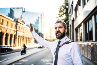 Hipster-Geschäftsmann, der auf der Straße in London steht und seine Hand hebt, um ein Taxi zu rufen. - HPIF30002