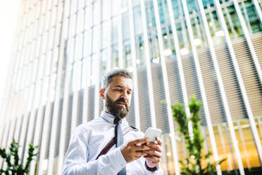 Low angle portrait of businessman with smartphone standing on the street in city in front of a building, text messaging. - HPIF30006