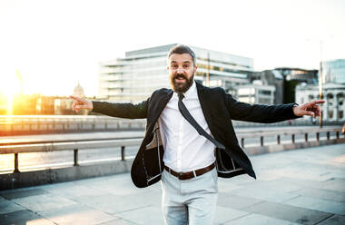 Cheerful hipster businessman dressed in white shirt and jacket walking on the bridge in the city, arms stretched. Success celebration concept. - HPIF30035