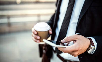 A close-up of an unrecognizable businessman with smartphone and coffee in a paper cup in the city, text messaging. Copy space. - HPIF30041