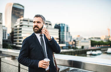 Seriöser Hipster mit Geschäftsmann mit Kaffee und Smartphone, der draußen in der Stadt steht und ein Telefonat führt. - HPIF30046