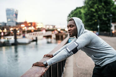 Young sporty black man runner with smartwatch, earphones and smartphone in an armband on the bridge in a city, resting. - HPIF30057