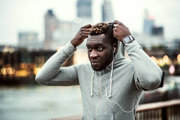 Junger sportlicher schwarzer Läufer mit Smartwatch, Kopfhörern und Smartphone in einer Armbinde auf der Brücke in einer Stadt, ruhend. Ein Mann, der eine Kapuze auf den Kopf setzt. - HPIF30058