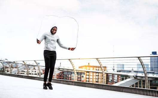 Nahaufnahme eines jungen aktiven schwarzen Sportlers, der mit einem Seil in einer Stadt springt und einen Kapuzenpulli trägt. - HPIF30066