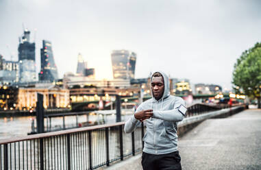 Junger sportlicher schwarzer Mann, der mit Smartwatch, Kopfhörern und Smartphone in einer Armbinde auf der Brücke in einer Londoner Stadt läuft und sich ausruht. - HPIF30083