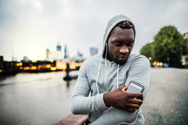 Junger sportlicher schwarzer Läufer mit Smartwatch, Kopfhörern und Smartphone in einer Armbinde auf der Brücke in einer Stadt, ruhend. - HPIF30084