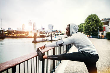 Young sporty black man runner with smartwatch, earphones and smartphone in an armband on the bridge in a city, stretching. - HPIF30085