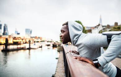 Young sporty black man runner with smartwatch, earphones and smartphone in an armband on the bridge in a city, resting. - HPIF30086