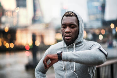 Young sporty black man runner with smartwatch, earphones and smartphone in an armband on the bridge in a London city, resting and measuring time. - HPIF30088