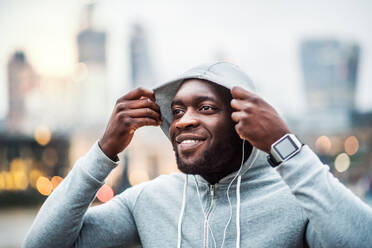 Young sporty black runner with smartwatch and earphones on the bridge in a London city, resting. A man putting a hood on head. - HPIF30089