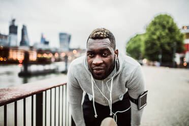 Junger sportlicher schwarzer Mann Läufer mit Smartwatch, Kopfhörer und Smartphone in einer Armbinde auf der Brücke in einer Stadt, ruhend. - HPIF30092