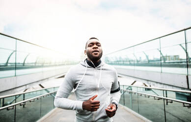 Junger sportlicher schwarzer Mann mit Smartwatch und Kopfhörern läuft auf der Brücke draußen in einer Stadt. - HPIF30094