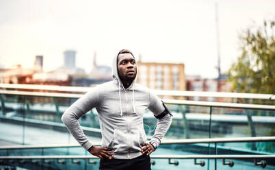 Junger sportlicher schwarzer Mann Läufer mit Smartwatch, Kopfhörer und Smartphone in einer Armbinde auf der Brücke in einer Stadt, ruhend. - HPIF30096