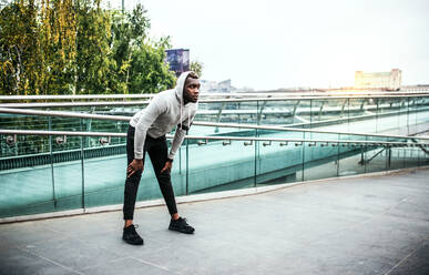 Junger sportlicher schwarzer Mann Läufer mit Kopfhörer und Smartphone in einer Armbinde auf der Brücke in einer Stadt, ruhend. - HPIF30101