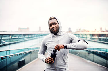 Ein junger sportlicher schwarzer Mann läuft mit einer intelligenten Uhr und einer Wasserflasche in einer Stadt. - HPIF30104