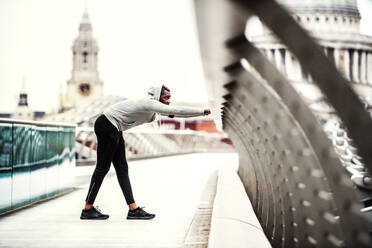 Junger sportlicher schwarzer Mann, der auf einer Brücke in einer Stadt läuft und sich streckt. - HPIF30106