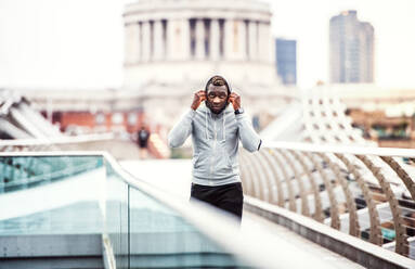 Junger sportlicher schwarzer Läufer mit Smartwatch, Kopfhörern und Smartphone in einer Armbinde auf der Brücke in einer Stadt, ruhend. - HPIF30107