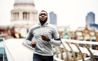 Junger sportlicher schwarzer Mann mit Smartwatch und Kopfhörern läuft auf der Brücke draußen in einer Stadt. - HPIF30108