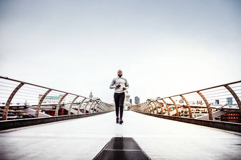 Junger sportlicher schwarzer Mann mit Smartwatch und Kopfhörern läuft auf der Brücke draußen in einer Stadt. - HPIF30115