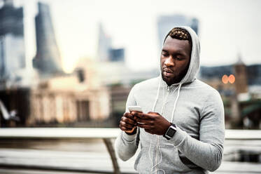 Young sporty black man runner with smartwatch, earphones and smart phone on the bridge in a city, resting. Copy space. - HPIF30118