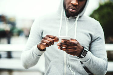 Unrecognizable young sporty black man runner with smartwatch, earphones and smart phone on the bridge in a city, resting. Copy space. - HPIF30119