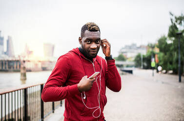 Junger sportlicher schwarzer Mann, der mit Kopfhörern, Smartwatch und Smartphone auf einer Brücke in einer Stadt läuft und Musik hört. - HPIF30144