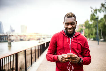 Junger sportlicher schwarzer Mann, der mit Kopfhörern, Smartwatch und Smartphone auf einer Brücke in einer Stadt läuft und Musik hört. - HPIF30145