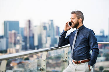A portrait of businessman with smartphone standing against London rooftop view panorama, making a phone call. Copy space. - HPIF30192