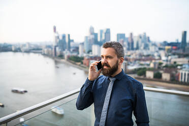 Porträt eines Geschäftsmannes mit Smartphone, der vor dem Panorama einer Londoner Dachterrasse steht und telefoniert. Kopierraum. - HPIF30200