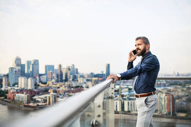 A portrait of businessman with smartphone standing against London rooftop view panorama, making a phone call. Copy space. - HPIF30201