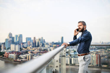 Porträt eines Geschäftsmannes mit Smartphone, der vor dem Panorama einer Londoner Dachterrasse steht und telefoniert. Kopierraum. - HPIF30201