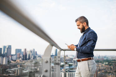 Porträt eines Geschäftsmannes mit Smartphone, der vor dem Panorama der Londoner Dachterrasse steht und eine SMS schreibt. - HPIF30202