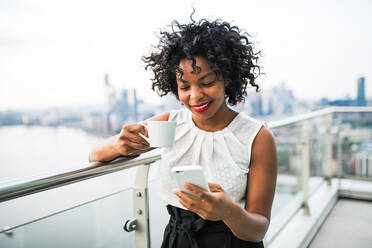 A businesswoman with coffee and smartphone standing against London view panorama, text messaging. - HPIF30219